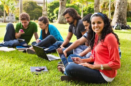Five students sat on grass studying, one of whom is looking into the camera